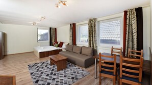 A modern living room with a sofa, a coffee table, and a dining area with chairs. There is a window with curtains, a radiator, and a bed in the background. - APLEND Bed & Breakfast Júlia (Vysoké Tatry)