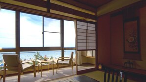 A room with tatami flooring, a low table, and chairs. Large windows provide a view of the beach and ocean. - Hotel Ichiboukan (Kyotango)