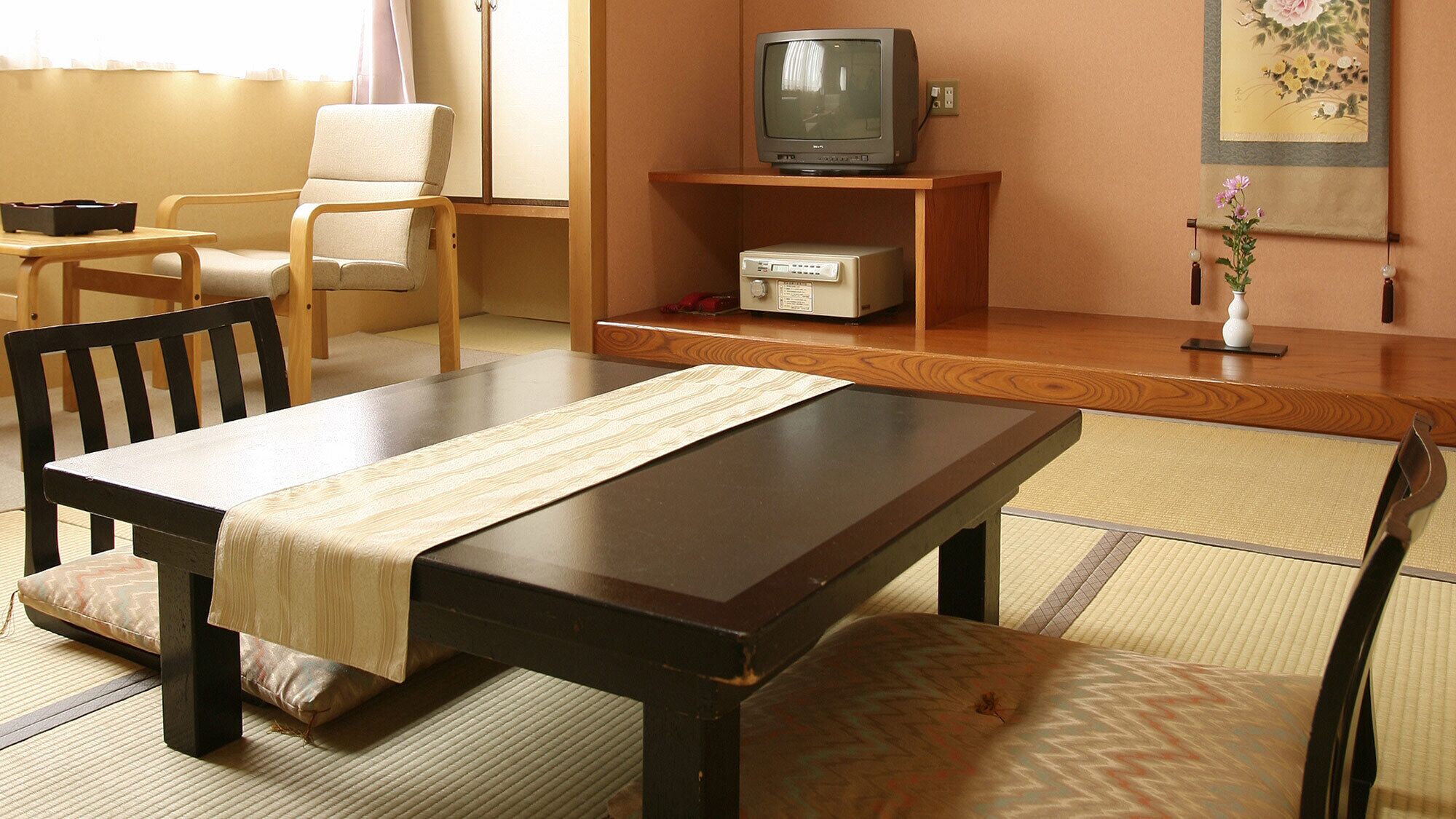 A traditional Japanese room with tatami flooring, a low wooden table, a television on a shelf, and a painting on the wall.