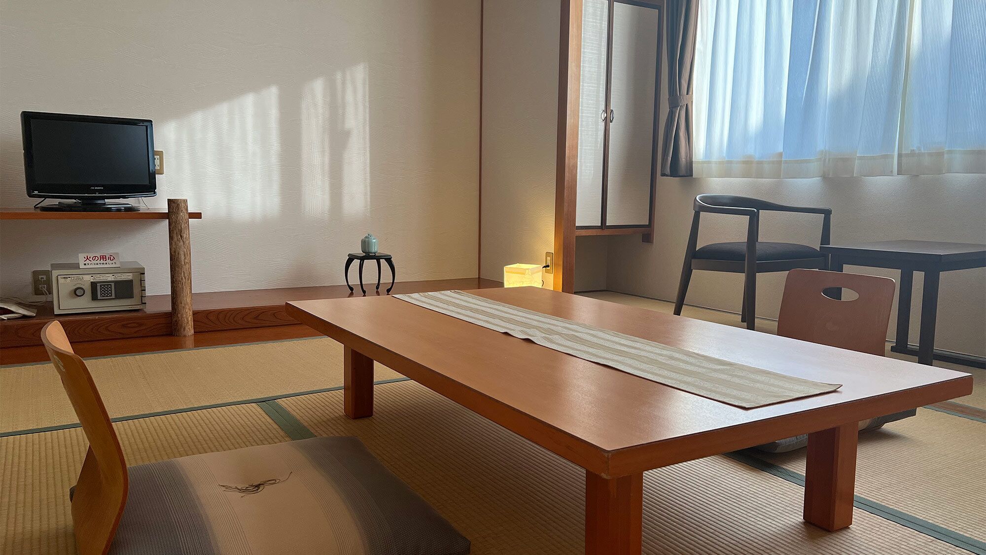 A traditional Japanese room with tatami flooring, a low table, a television on a shelf, and a window with sheer curtains.