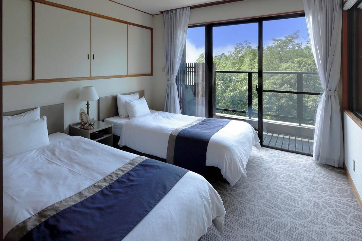 A hotel room with two beds, a nightstand, a lamp, and a large window with curtains.