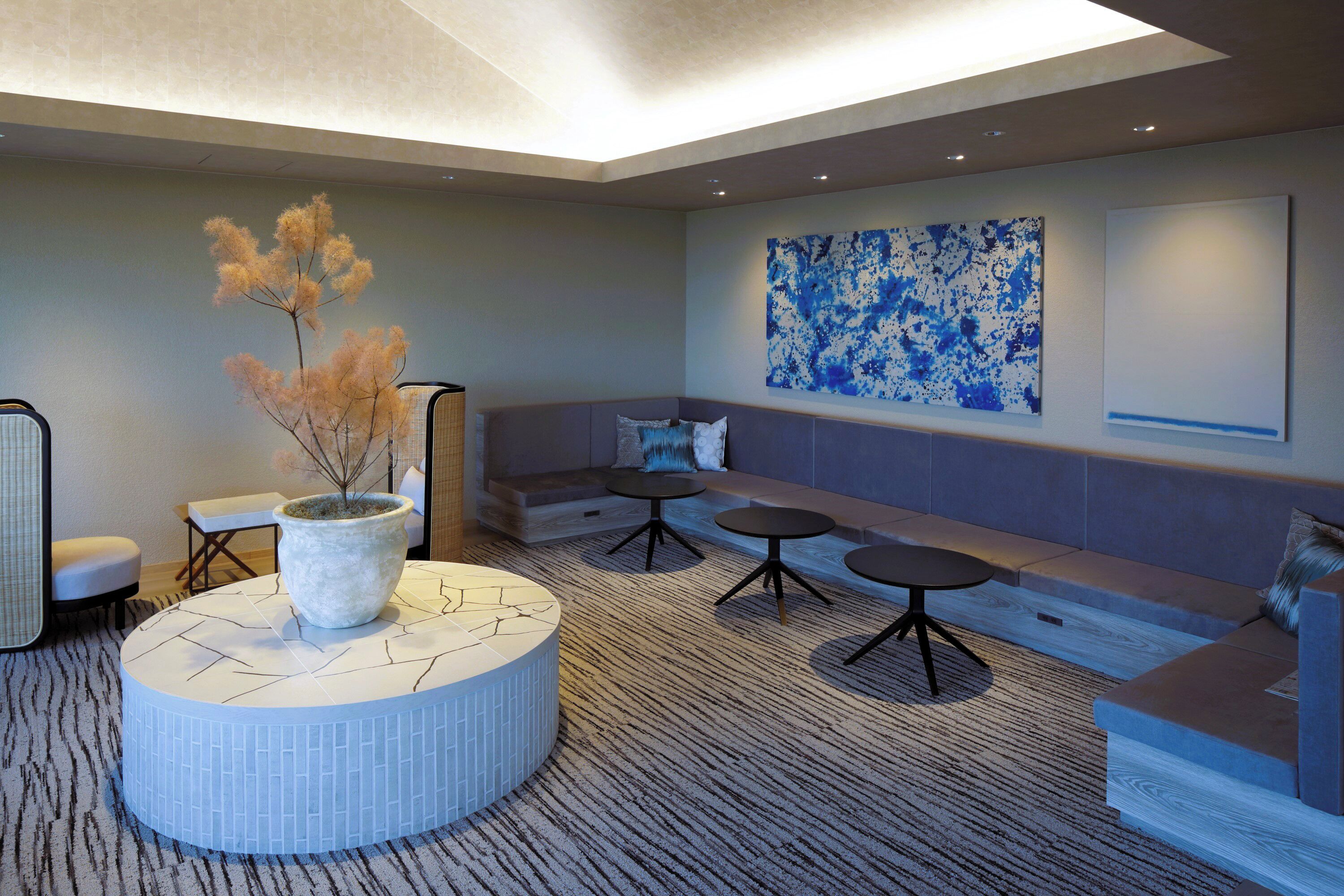 A modern lounge area with a round seating arrangement, a large abstract painting, and a potted plant.