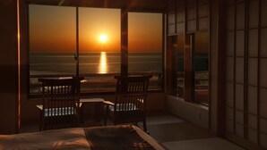 Quarto com cama, duas cadeiras, uma mesinha e vista para o oceano ao pôr do sol.