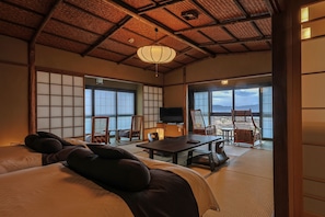 Una habitación tradicional japonesa con suelo de tatami, una cama grande, un televisor y vistas a la montaña a través de amplios ventanales.