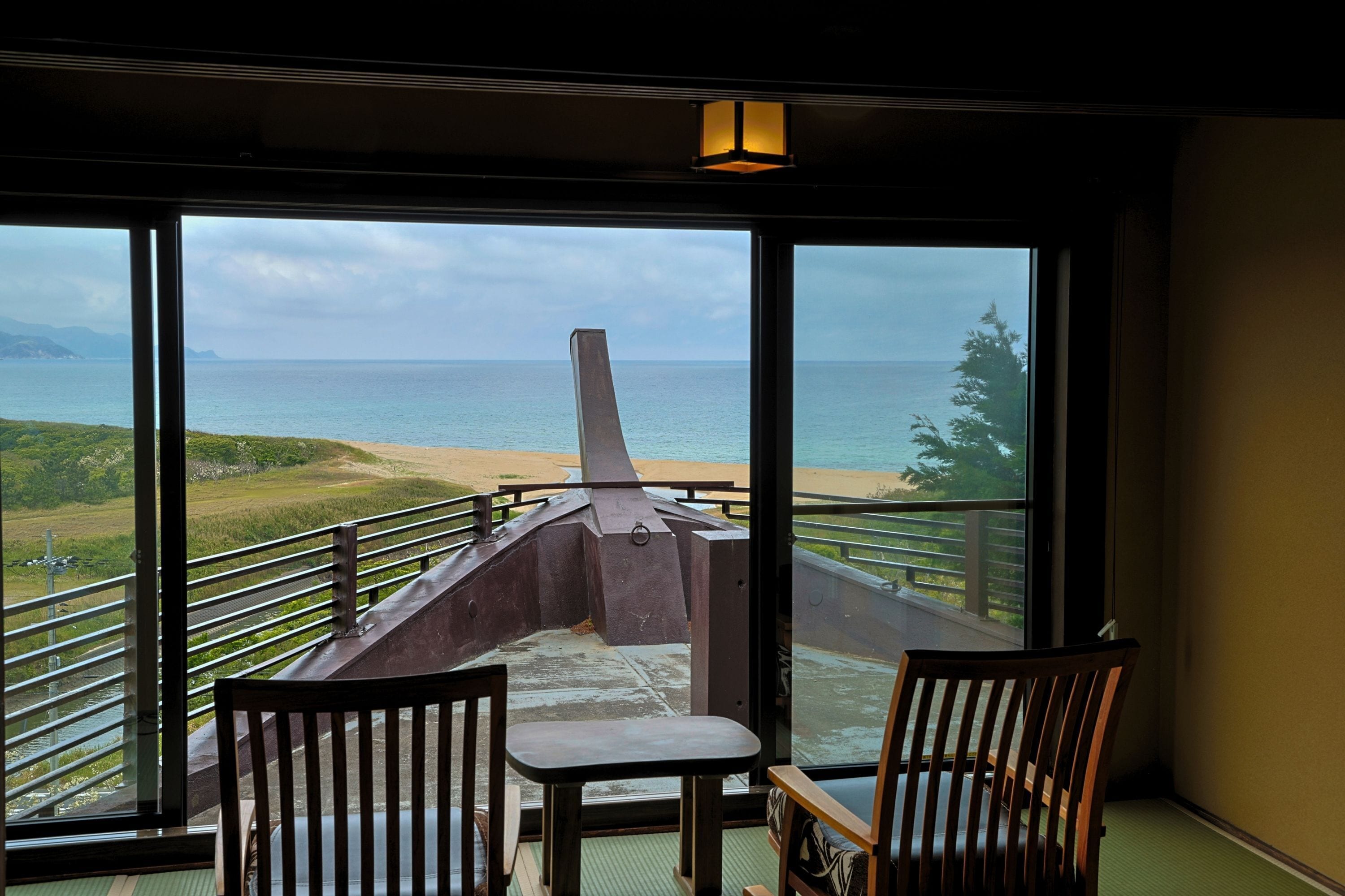 A room with a large window overlooking a beach and ocean. There are two wooden chairs and a small table.
