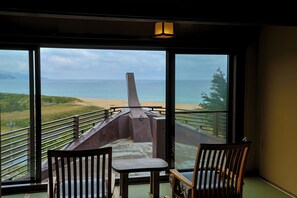 Een kamer met een groot raam met uitzicht op een strand en de oceaan. Er staan twee houten stoelen en een kleine tafel.