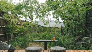 A wooden deck with a round table and two woven stools, surrounded by lush greenery and bamboo structures. -  Ujo Soan (Kyotango)