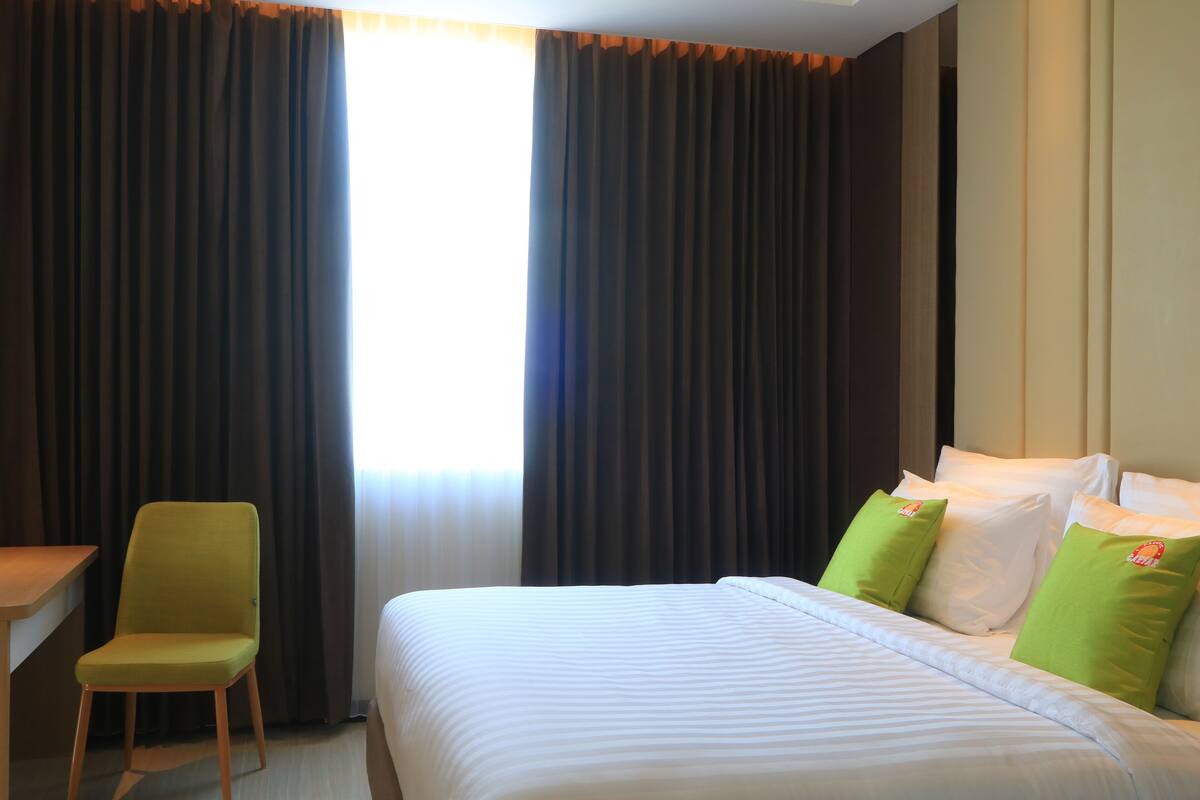 Una habitación de hotel con una cama, almohadas verdes, una silla de madera y cortinas.