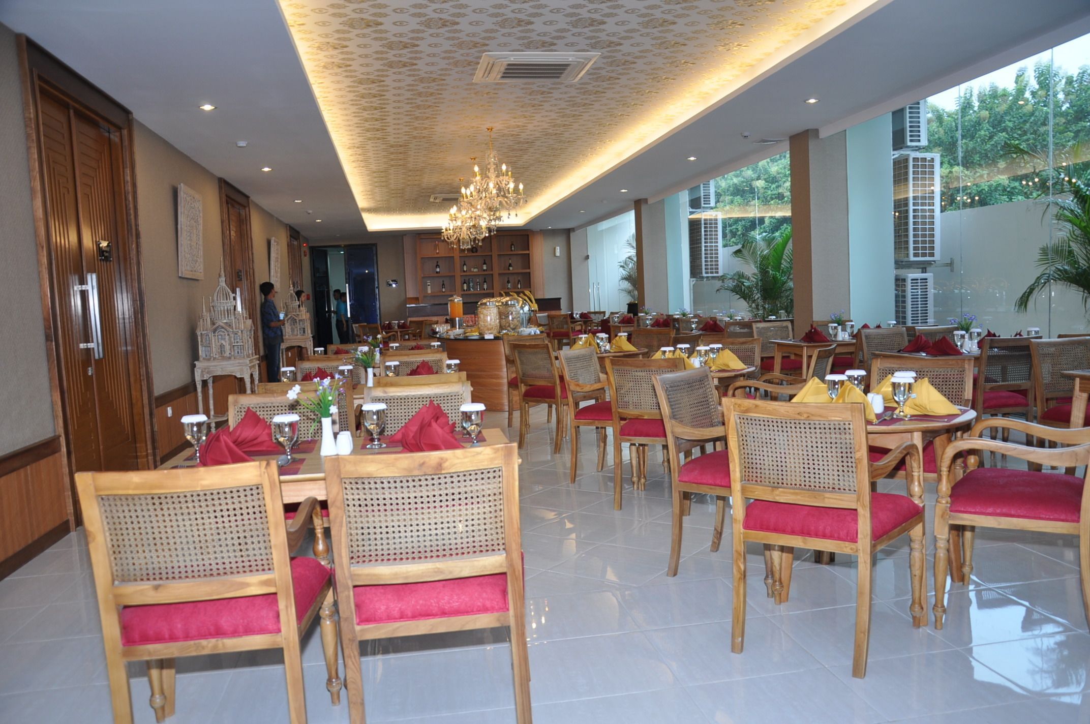 A spacious dining area with wooden tables and chairs, a chandelier, and large windows offering a view of greenery outside.