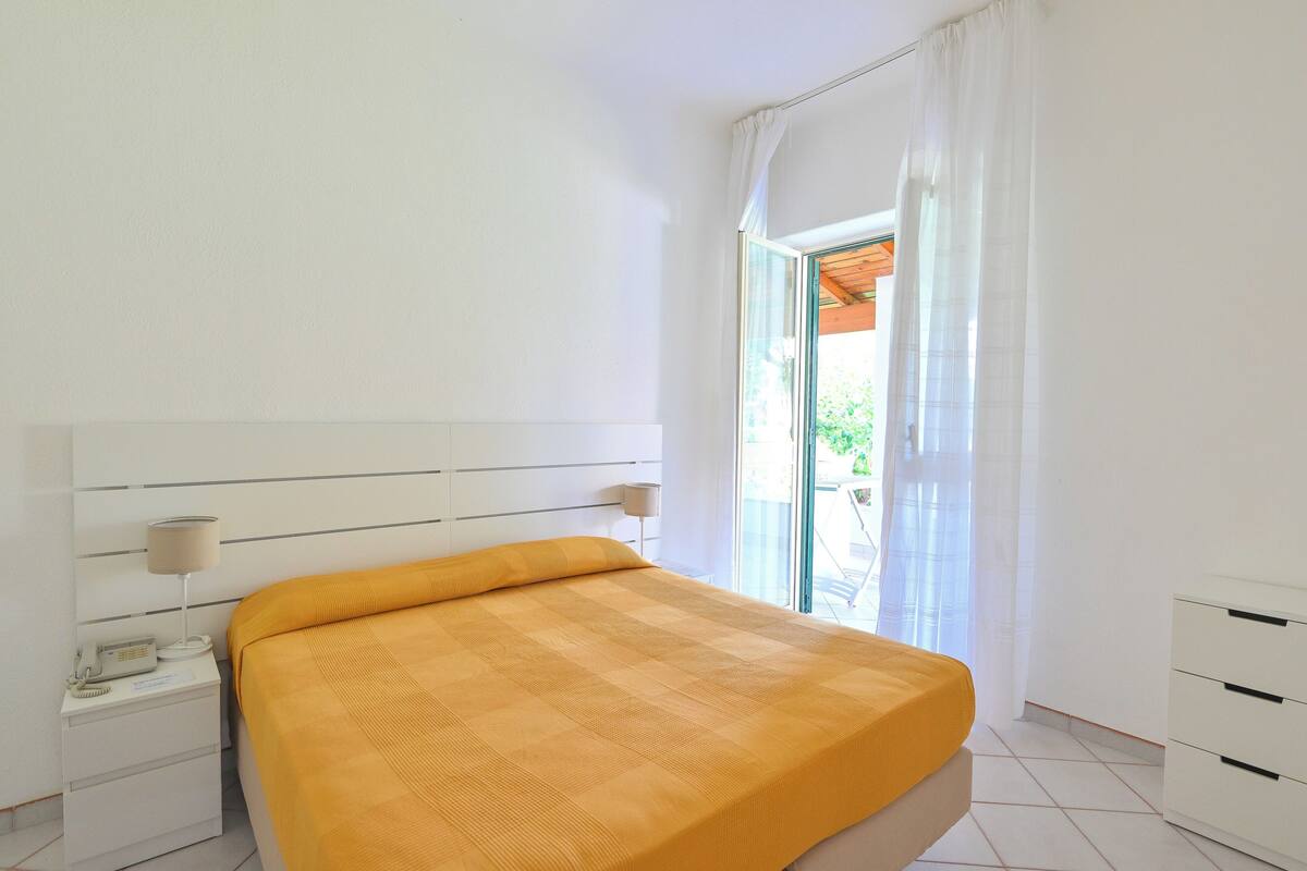 Un dormitorio con cama, mesita de noche con lámpara, mesita auxiliar y ventana con cortinas.