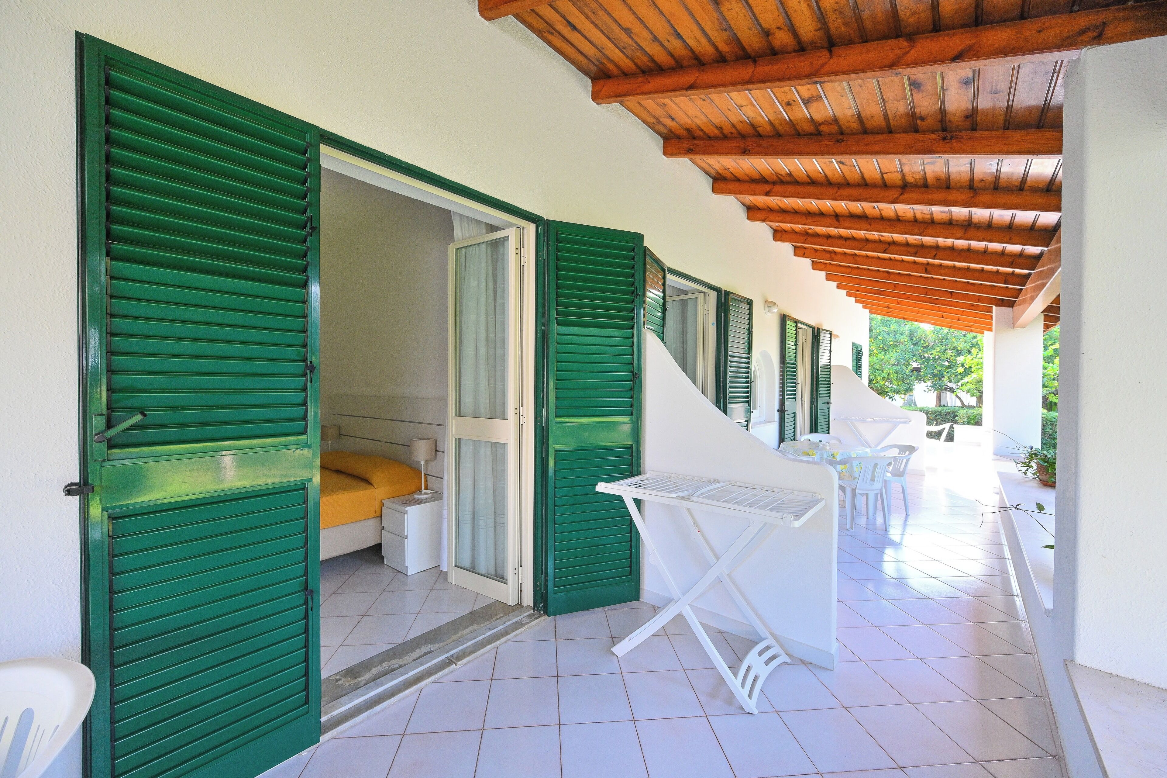 Un espacio exterior techado con suelo de baldosas, una mesa plegable blanca y contraventanas verdes en las paredes.