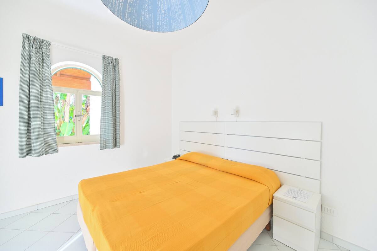 Un dormitorio con cama, ventana con cortinas, cabecera blanca y mesita de noche.