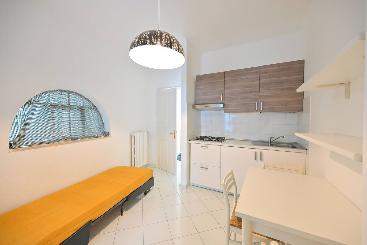 Cocina compacta con muebles blancos, estufa y lavabo. Hay un banco y una silla en la habitación.