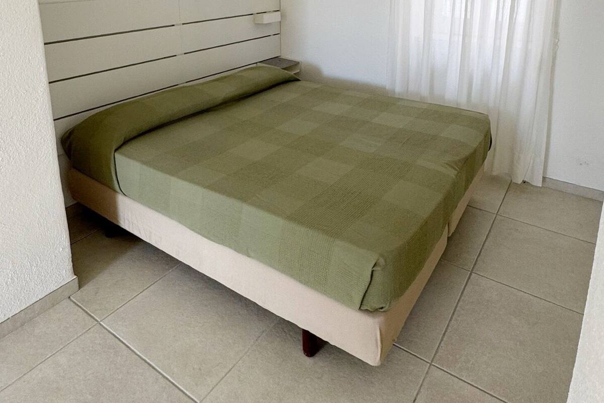 Habitación individual con una cabecera, un equipo de aire acondicionado en la pared y una ventana con cortinas blancas.