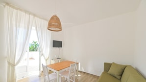 A room with a wooden dining table, white chairs, a green sofa, a TV on the wall, and a wicker pendant light. - Residence San Domenico (Forio)