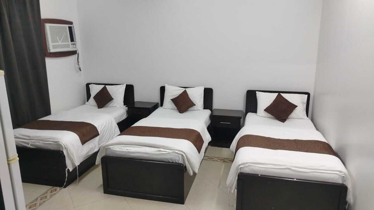 A hotel room with three single beds, each with a pillow and a bedside table.