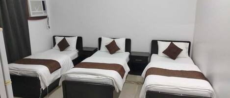 A hotel room with three single beds, each with a pillow and a bedside table.