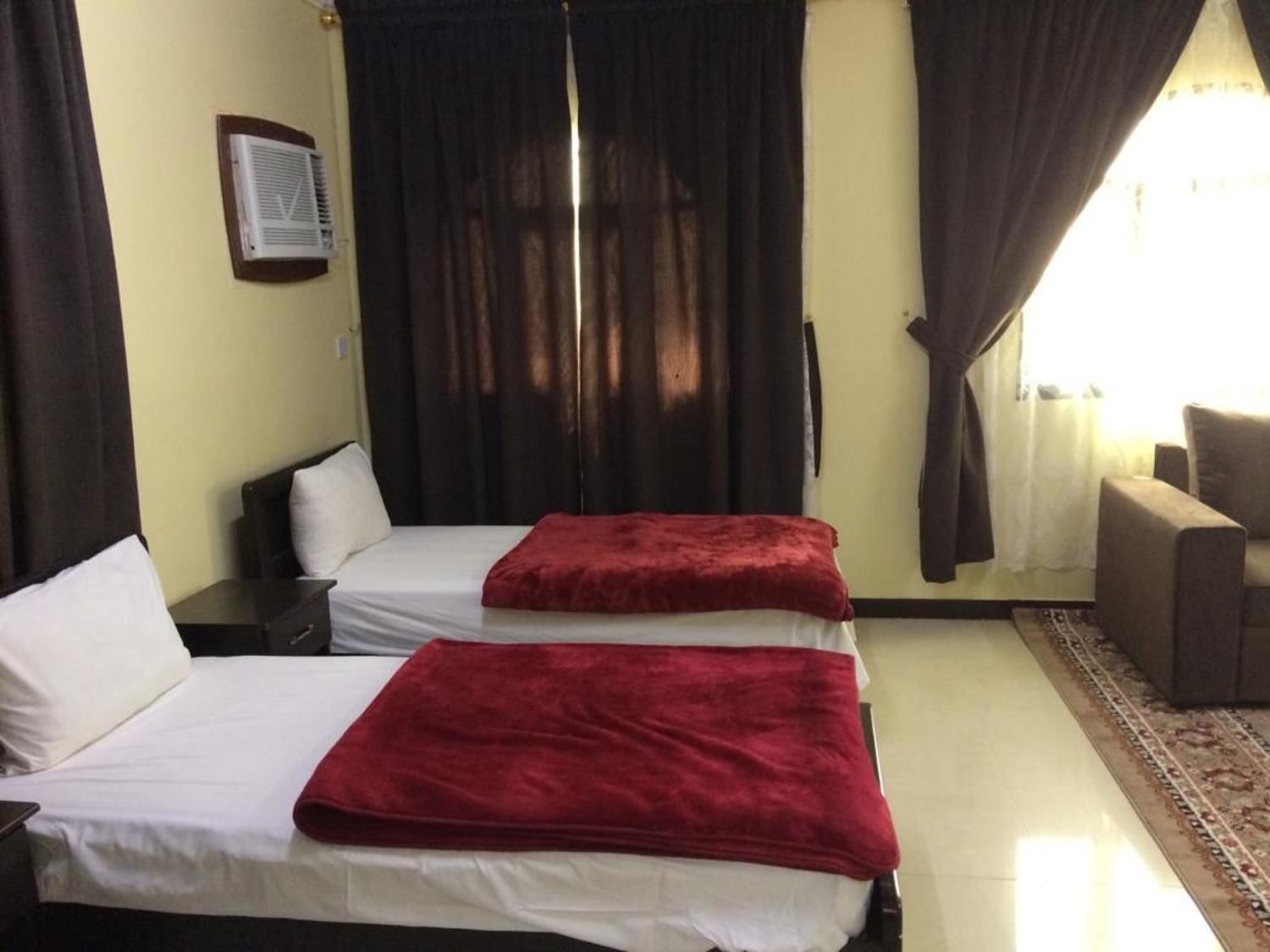A hotel room with two beds, a red blanket on each, a small table, a chair, and a window with curtains.