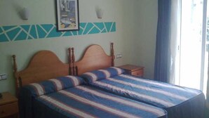 Um quarto com cama de madeira, roupa de cama listrada, uma pequena mesa de cabeceira, um criado-mudo, uma luminária fixada na parede e uma janela com cortinas.