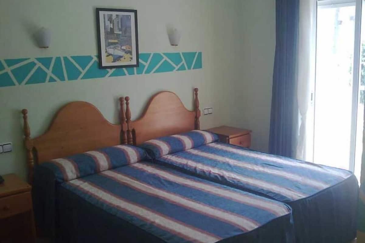 Un dormitorio con cama de madera, ropa de cama a rayas, una mesita de noche pequeña, una lámpara de pared y una ventana con cortinas.