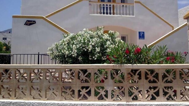 Um edifício de dois andares com fachada branca, varanda com grade e uma pequena janela. Há uma placa com o nome 'Agadir' e uma placa de estacionamento com um 'P' no edifício.