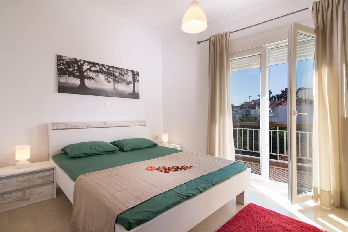 Un dormitorio con cama, ropa de cama verde, una mesita de noche con lámpara, un cuadro en la pared y un balcón con cortinas.