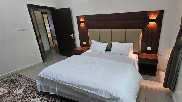 A hotel room with a large bed, a wooden headboard with integrated lighting, a nightstand, and a bedside table. - Al Eairy Serviced Apartments Company  (Buraydah)