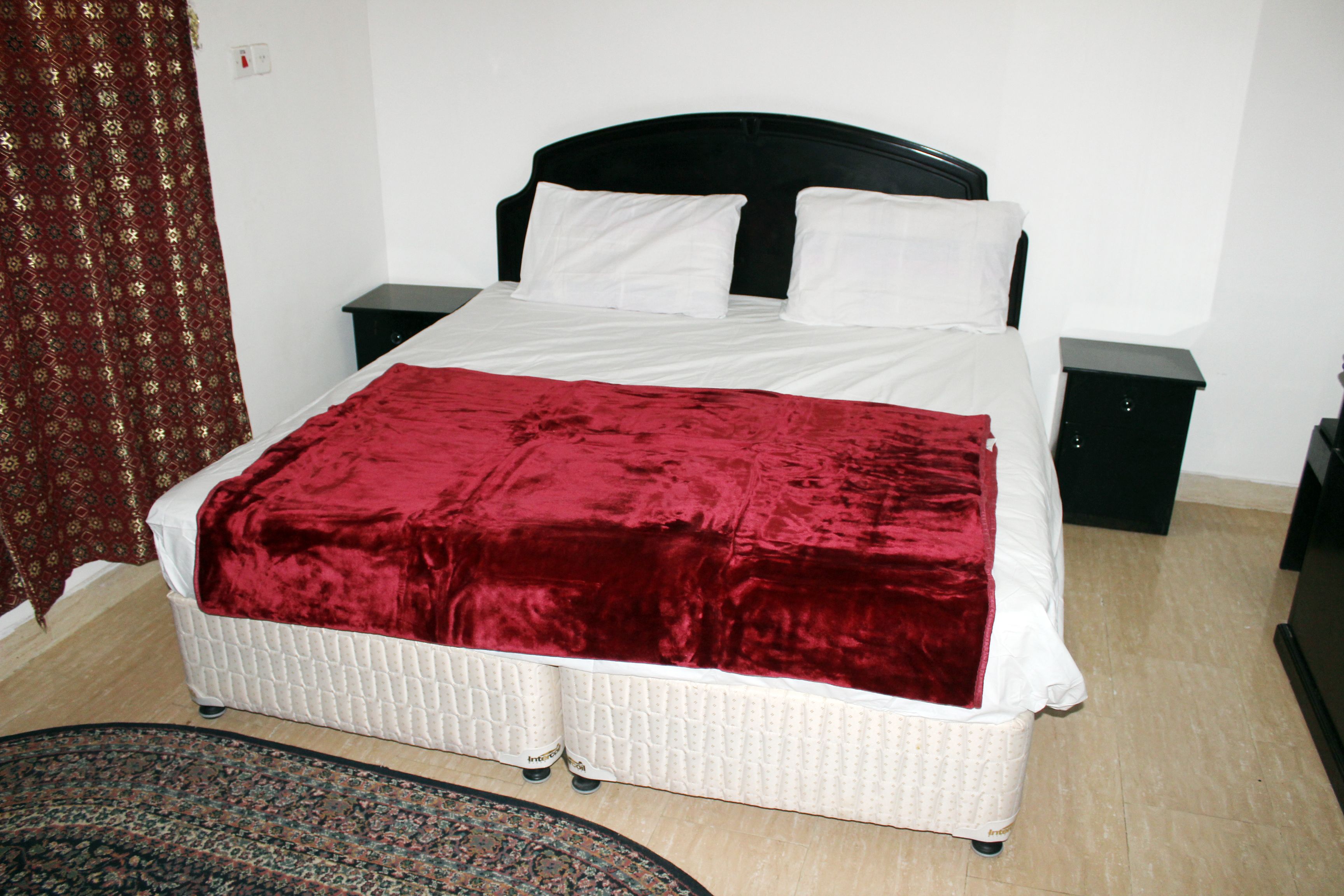 A neatly made bed with a red blanket, white pillows, and a black headboard. There are two bedside tables, one on each side of the bed.