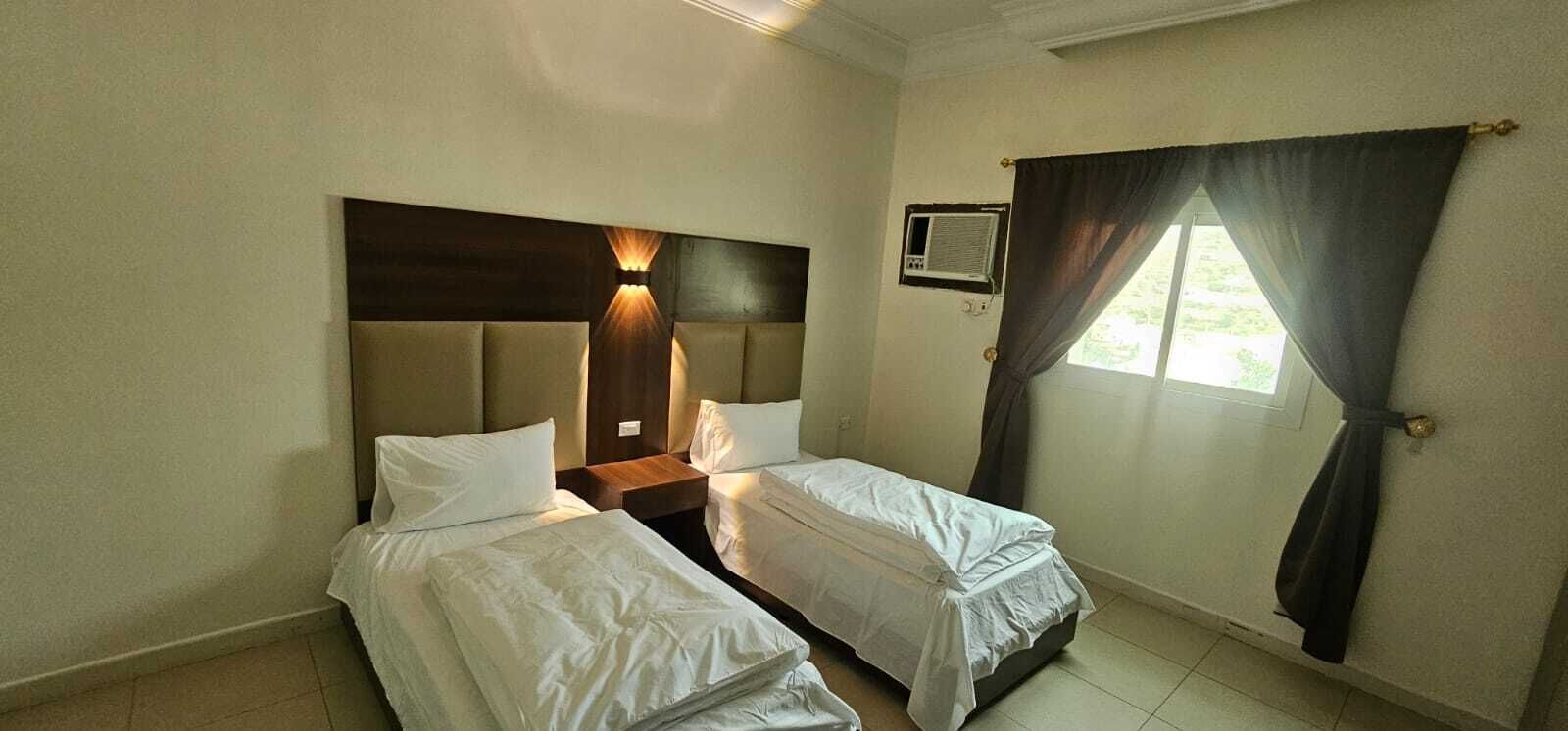 A hotel room with two single beds, a wooden headboard, a small air conditioning unit, and a window with dark curtains.
