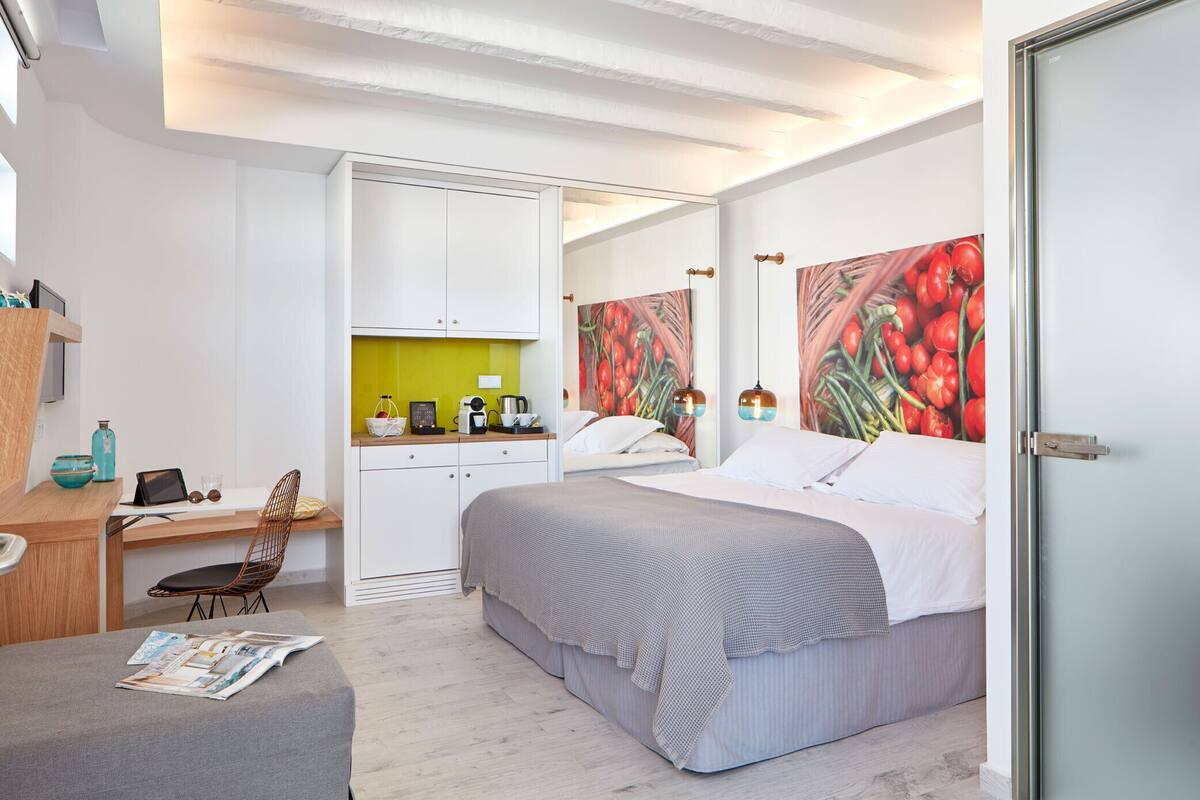 A modern bedroom with a bed, a desk, and a chair. There is a window, a door, and a wall with a fruit painting.
