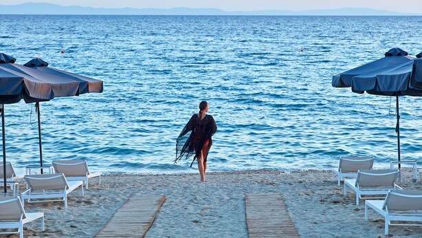 En strand med en person, der går mod havet, omgivet af strandstole og parasoller.
