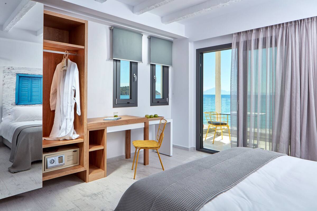 A modern hotel room with a bed, a desk, a chair, a wardrobe, and a balcony with a view of the sea.