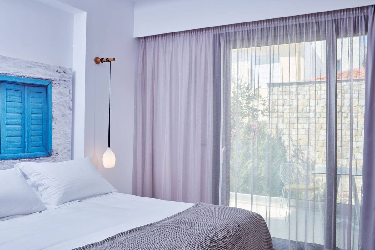 A bedroom with a bed, a window with curtains, a wall with a blue shutter, and a hanging light fixture.