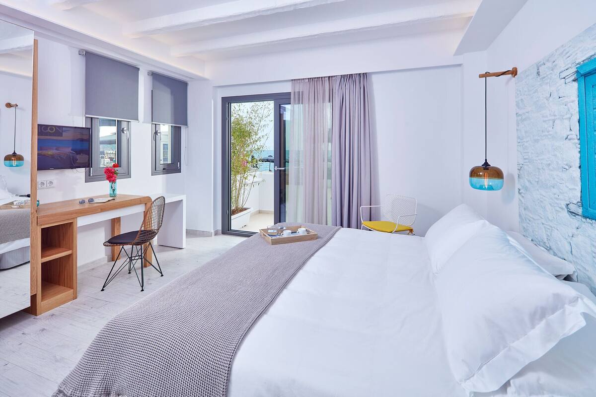 A modern hotel room with a bed, a desk, and a chair. There is a sliding glass door leading to an outdoor area.