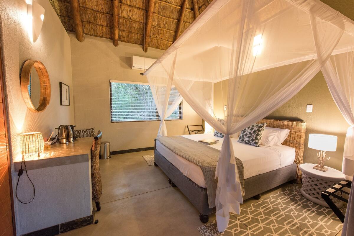 A hotel room with a bed under a canopy, a bedside table with a lamp, a small round table with a lamp, a window with blinds, and a wall-mounted air conditioner.
