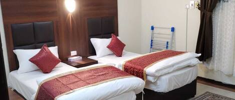 A hotel room with two beds, each with a red accent pillow, a wooden headboard, and a bedside table. There is a large patterned rug on the floor and an air conditioning unit on the wall.