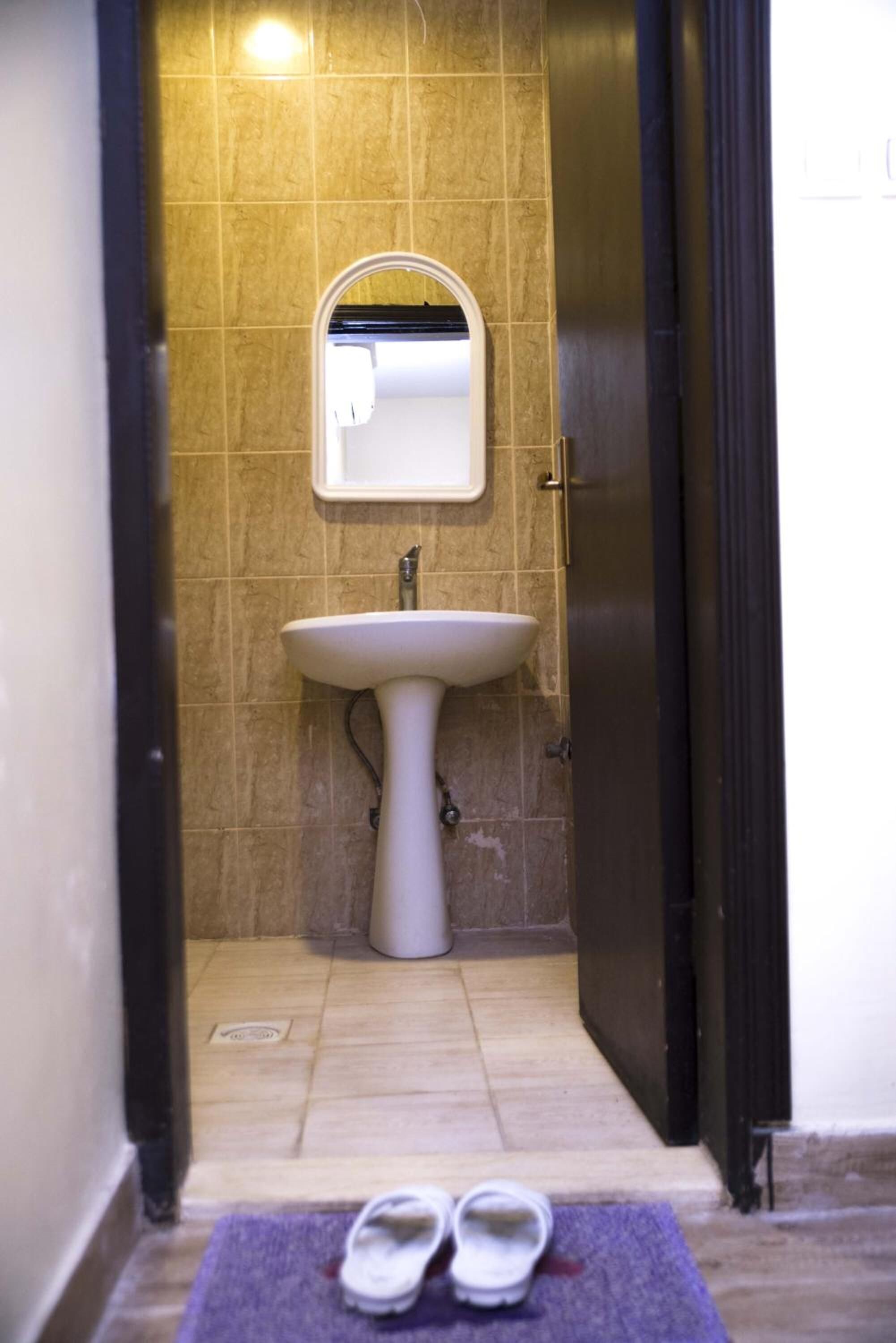 A narrow bathroom with a pedestal sink, a mirror, and a pair of slippers on a mat.