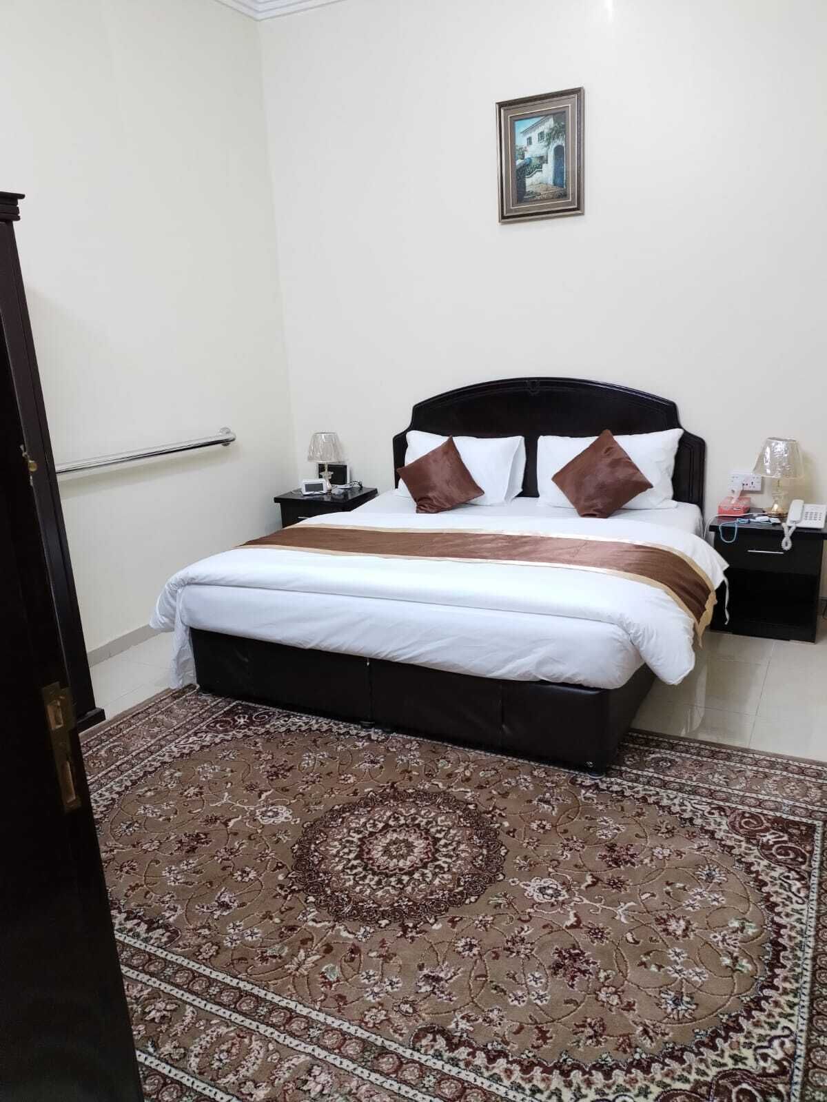 Una cama bien hecha con una colcha marrón y blanca, un cabecero oscuro y una alfombra estampada en el suelo.