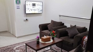 Een woonkamer met een bruine zithoek, een salontafel met zakdoosjes en bloemen, een kamerbreed tapijt, een flatscreen televisie aan de muur en een ingelijste foto boven de bank.