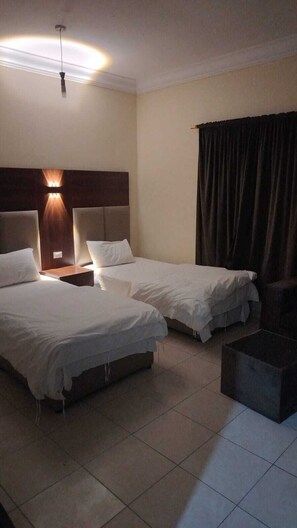 Habitación de hotel con dos camas, una mesita de noche, una lámpara, una cortina y una pequeña mesa.