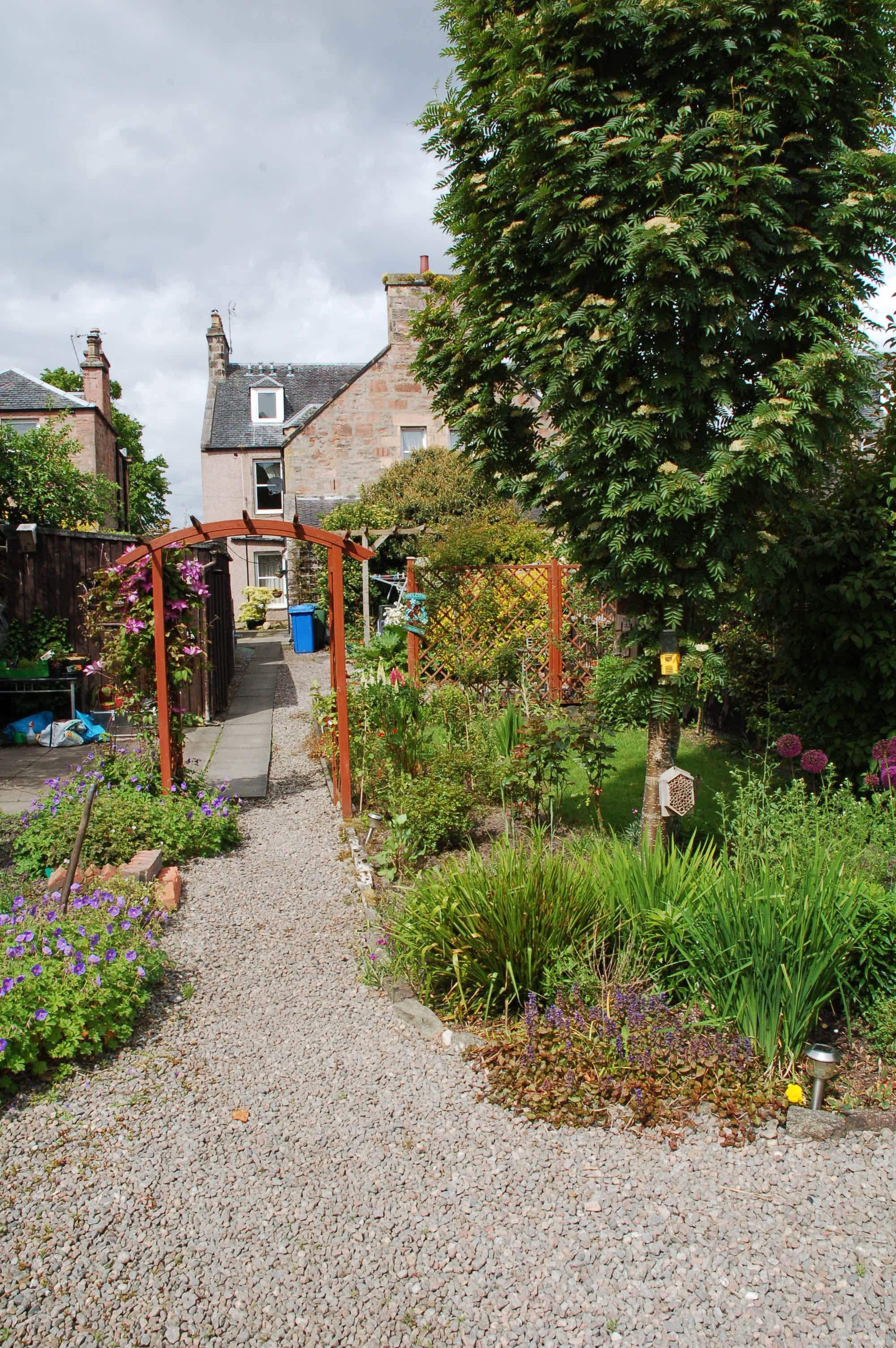 A garden pathway leading to a house with a wooden pergola and various plants.
