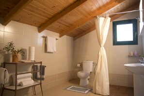 A bathroom with a wooden ceiling, a white toilet, a sink, a shower curtain, and a towel rack with folded towels. - Agrotospita Country Houses (Nafplio)