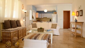 A living room with a sofa, a coffee table, and a small table with a lamp. There is a kitchen in the background with cabinets and a refrigerator. - Agrotospita Country Houses (Nafplio)