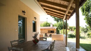 A covered patio with a wooden table and chairs, a bowl of fruit, and a view of a house with a red-tiled roof. - Agrotospita Country Houses (Nafplio)