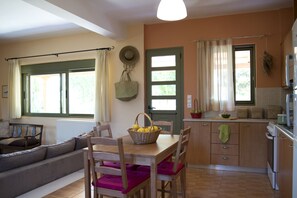 A cozy living room with a sofa, dining table, and chairs. A kitchen with a stove, sink, and cabinets is visible in the background.