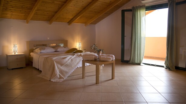 A bedroom with a wooden ceiling, a bed with white linens, a bedside table with pillows, and a window with curtains. - Agrotospita Country Houses (Nafplio)