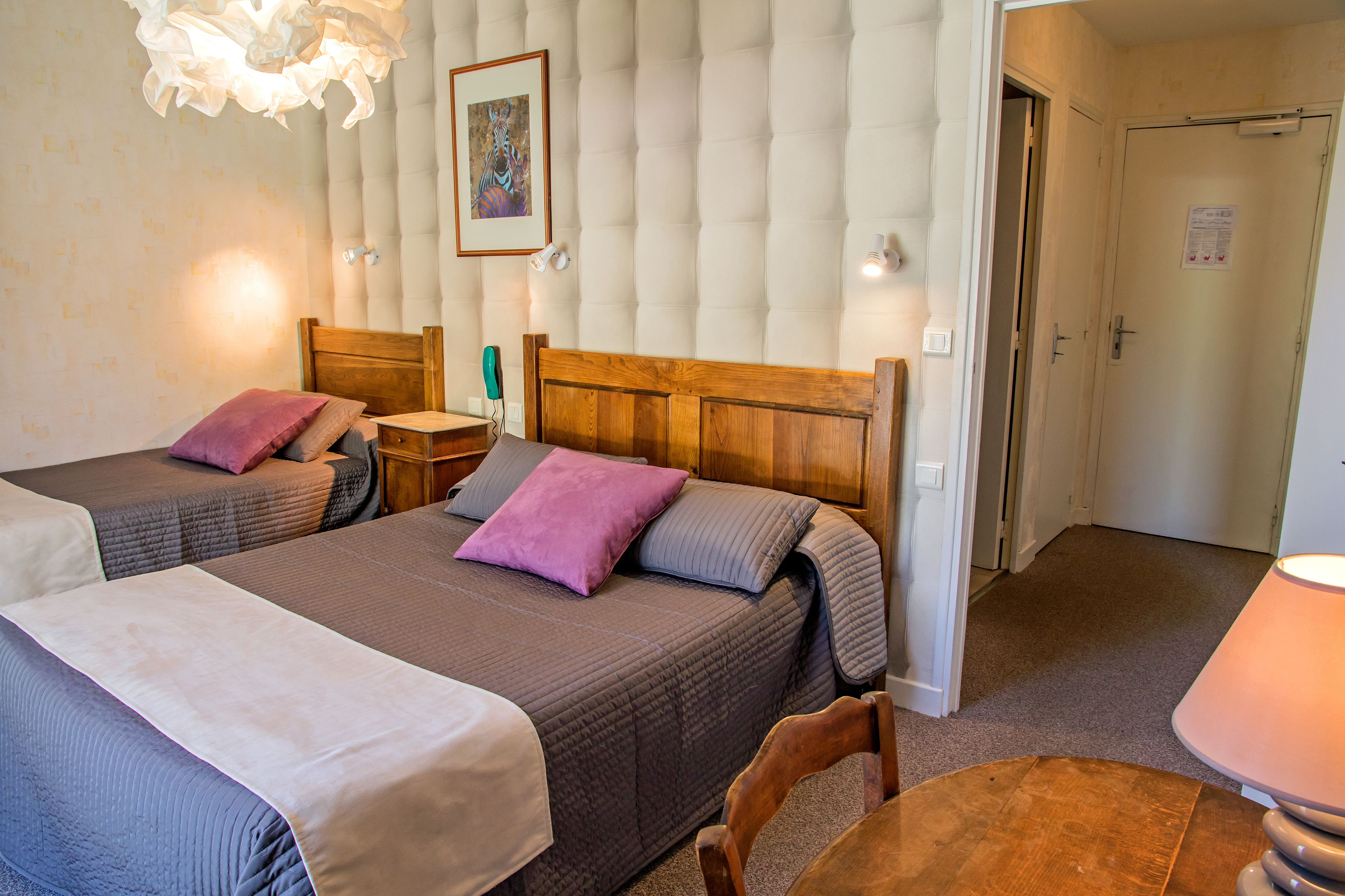 A hotel room with two beds, a wooden headboard, a bedside table, a lamp, and a door.