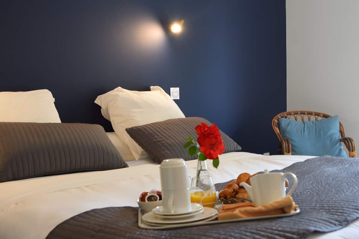 Un lit bien fait, avec des oreillers, un plateau de petit-déjeuner et un vase contenant une fleur rouge.