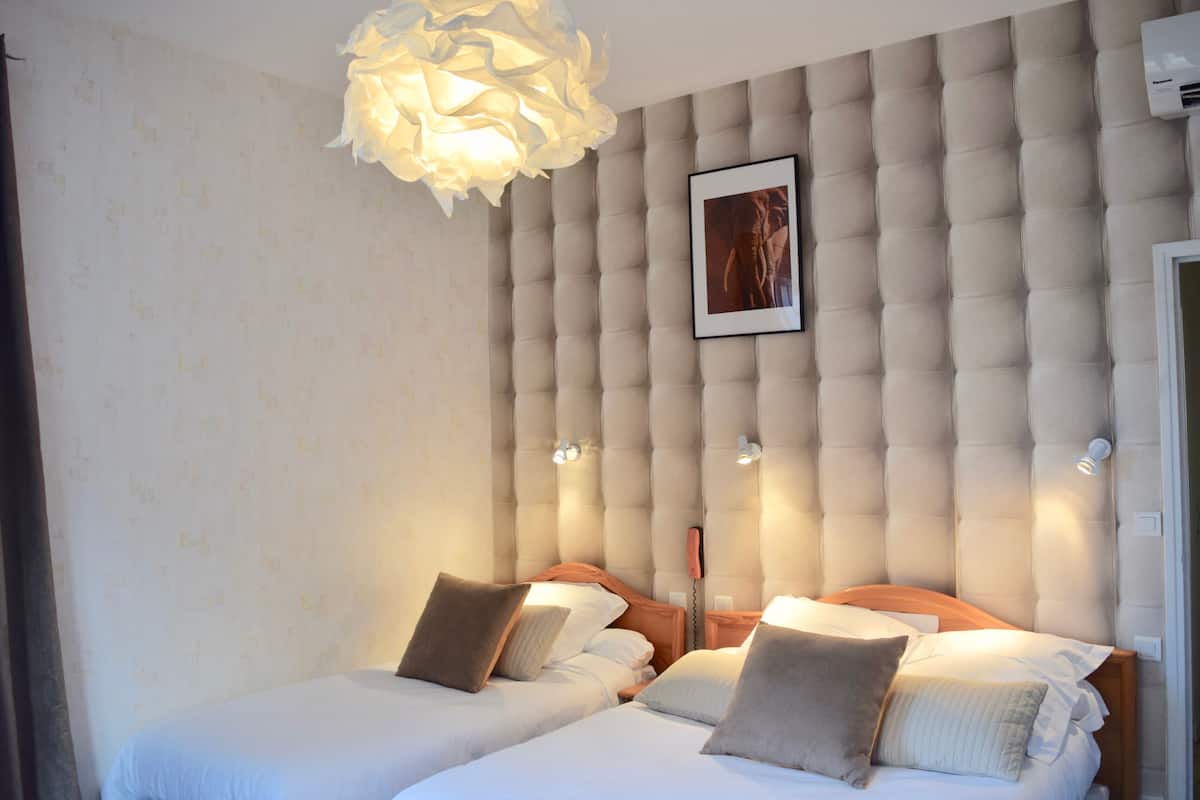 Une chambre d’hôtel avec deux lits, une tête de lit capitonnée, un luminaire suspendu et un tableau encadré au mur.