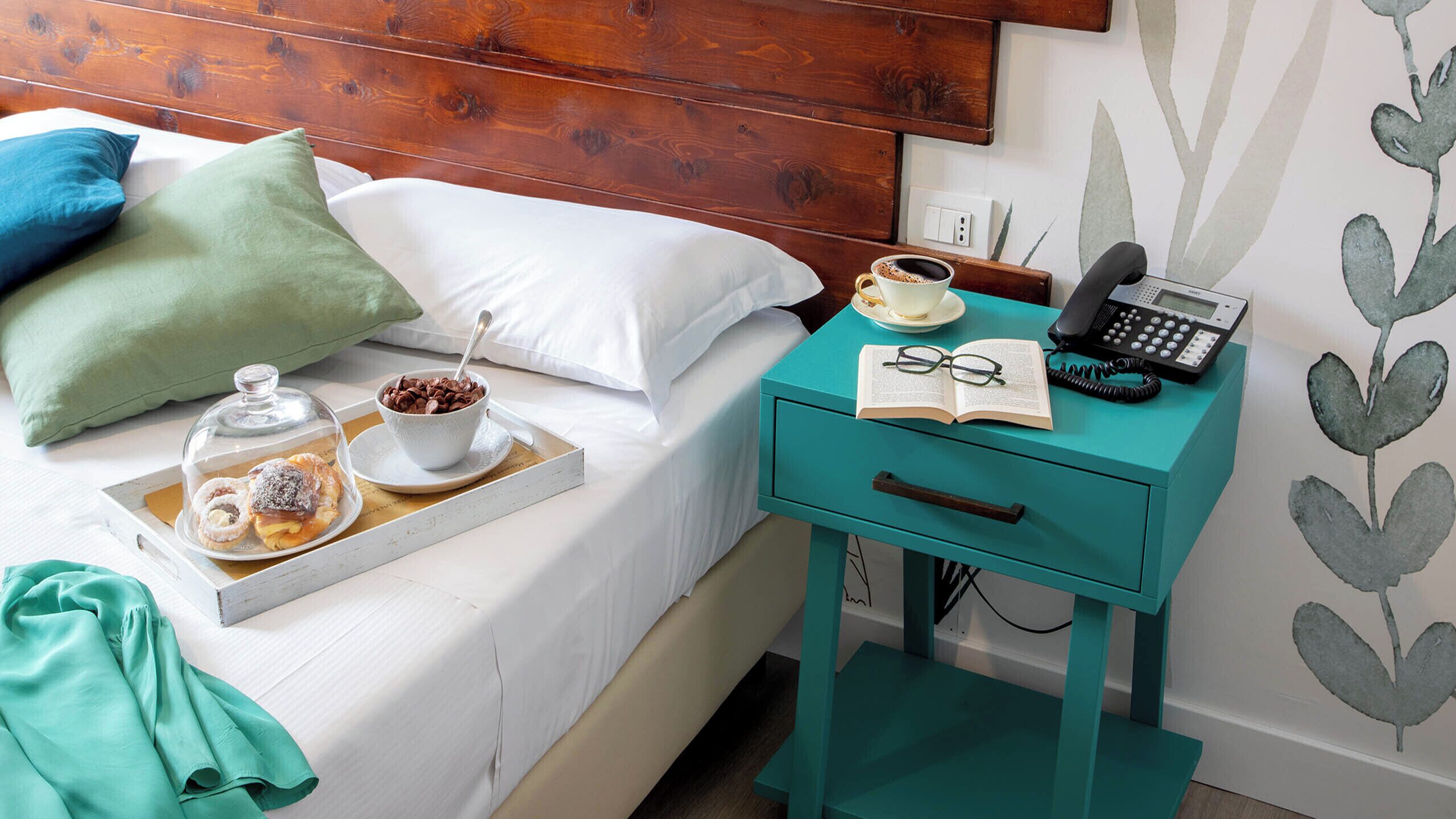 A neatly made bed with pillows, a tray with pastries and a bowl of chocolate, a teal nightstand with a book, glasses, and a phone.